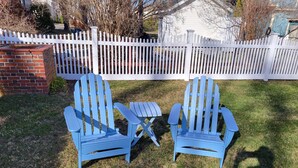 Two blue Adirondack chairs with a small table in a grassy yard, a white picket fence, and a brick structure in the background. - Cozy Cottage at Footbridge Beach - Great Fall and Spring Rates (Ogunquit)