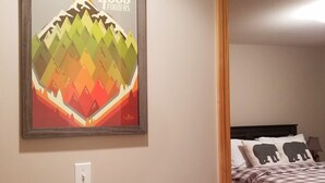 Une affiche encadrée accrochée au mur, un interrupteur et un lit recouvert d’une couverture à carreaux et orné de deux coussins à motifs d’éléphants.