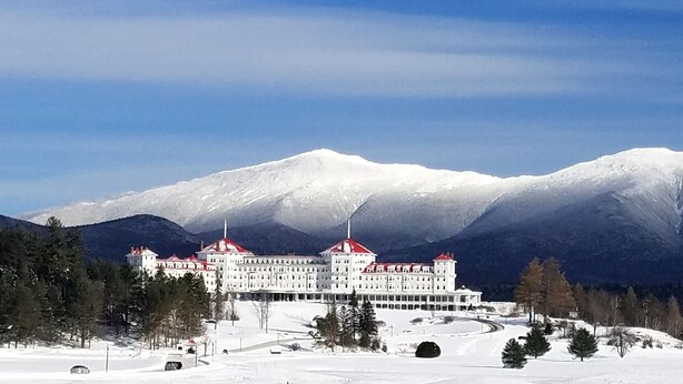 Un grand bâtiment blanc, surmonté de toits rouges, entouré de montagnes et d’arbres couverts de neige.