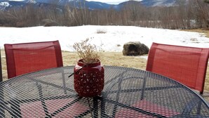 Une table ronde, ornée d’un vase rouge au centre, est entourée de chaises rouges. L’ensemble est placé devant un paysage enneigé, avec des montagnes en arrière-plan.
