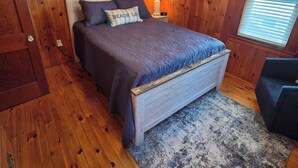 Une chambre avec un plafond en bois, un lit avec une tête de lit, une table de chevet avec une lampe, une fenêtre avec des stores et une chaise.