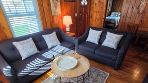 Une pièce avec deux canapés sombres, une table basse ronde en bois, un tapis à motifs, des murs en bois, une porte et un lit à l’arrière-plan.