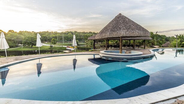 A swimming pool with a thatched-roof gazebo, surrounded by palm trees and a view of a golf course. - Rio das Garças Eco Resort (Sao Jose de Mipibu)