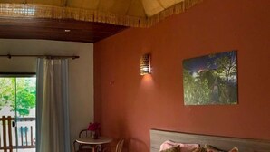 Une chambre à coucher avec un lit, des oreillers, un tableau, une petite table avec des chaises et une vue par la fenêtre.