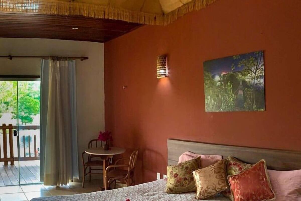 Quarto com cama, almofadas, um quadro, uma mesinha com cadeiras e vista pela janela.