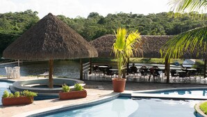 Un espace piscine agrémenté d’un parasol en chaume, de plantes en pot et de coins salon extérieurs situés au bord de l’eau.