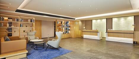 A modern hotel lobby with a reception desk, seating area with sofas and chairs, and a large bookshelf filled with books.