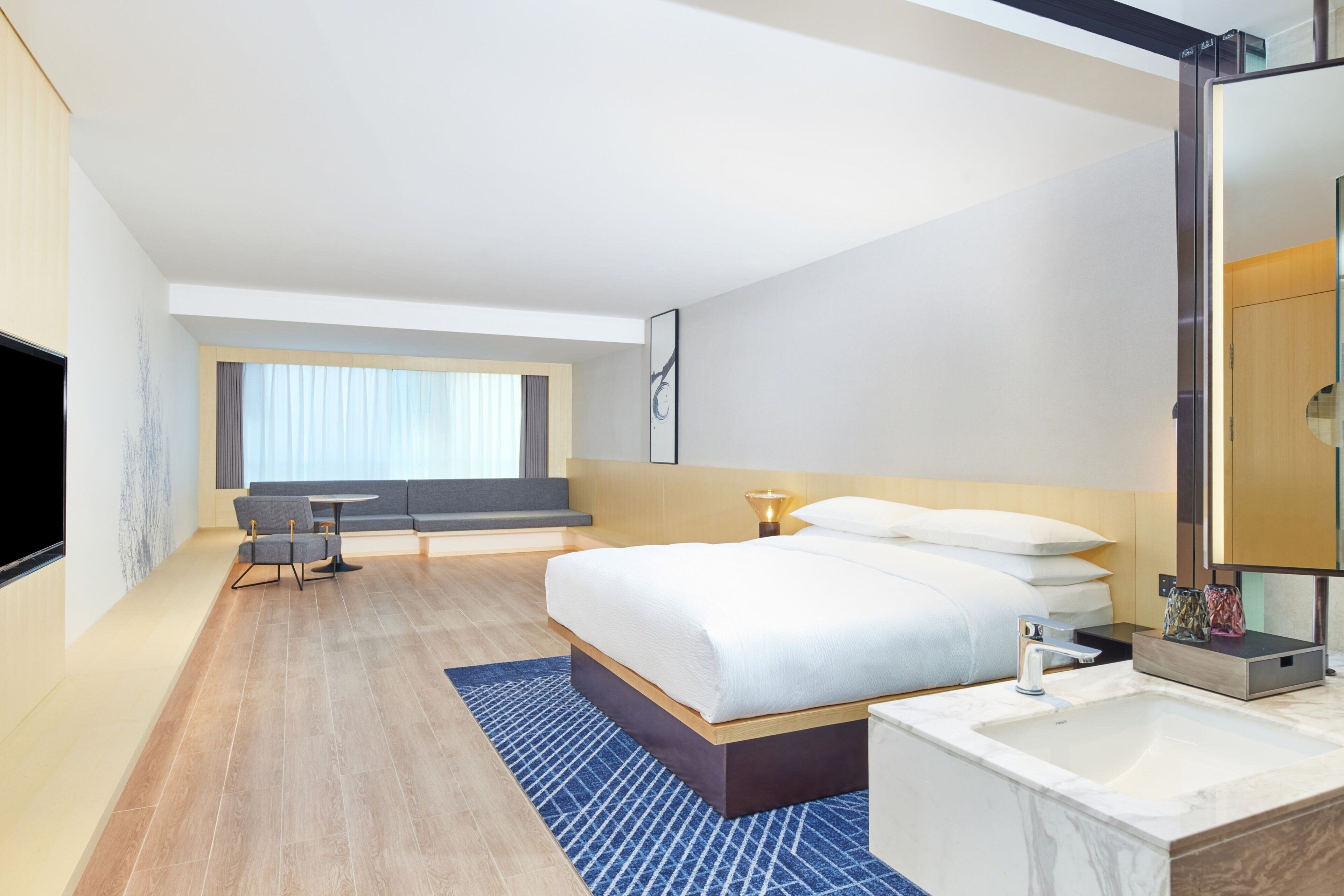 Una habitación de hotel moderna con una cama grande, una mesita de noche, televisión integrada, una ventana con cortinas y una zona de estar con sofá y sillones.