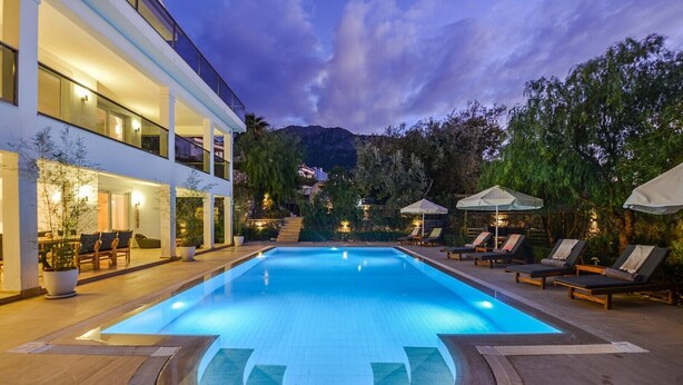 A swimming pool with lounge chairs and umbrellas, surrounded by a landscaped garden and a building with a balcony. - 5 bedroom luxury villa with private pool and garden in oludeniz (Fethiye)