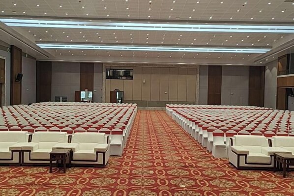 A large conference room with rows of white chairs and red carpets, equipped with a podium and a large screen.