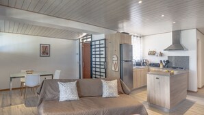 A modern living room with a sofa, a dining area with chairs, a kitchen with a stove and refrigerator, and a wooden ceiling.