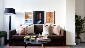 A living room with a brown sofa, three framed pictures on the wall, a black floor lamp, a small table with bowls and a book, and a potted plant. - Watford WD18 - Beautiful 1Bedroom/1Bathroom Flat with Free Parking & Free WIFI (Watford)