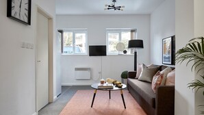 A modern living room with a sofa, coffee table, and a floor lamp. There are two windows with curtains, a wall-mounted TV, and a clock on the wall. - Watford WD18 - Beautiful 1Bedroom/1Bathroom Flat with Free Parking & Free WIFI (Watford)