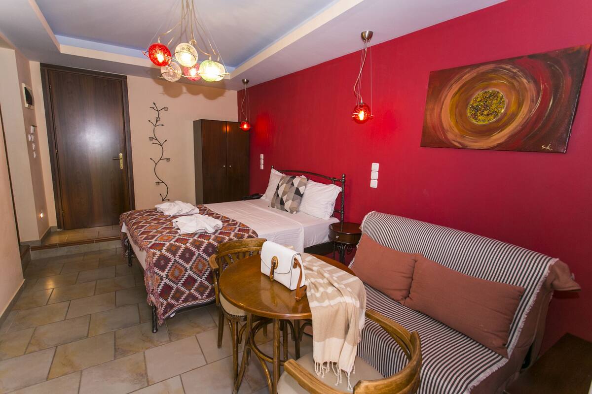 A room with a bed, a sofa, a dining table, and a chair. There is a painting on the wall and a door.