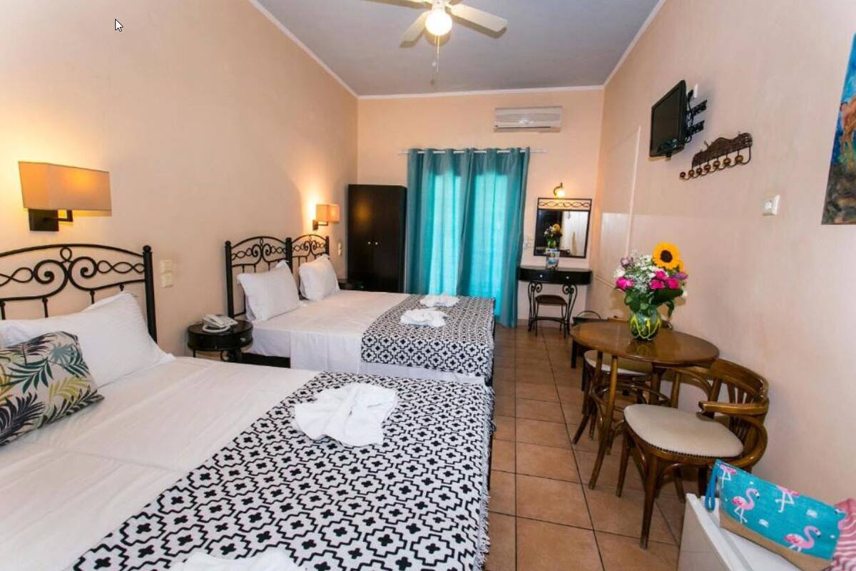 A hotel room with two beds, a desk, a chair, a table with a vase of flowers, and a ceiling fan.