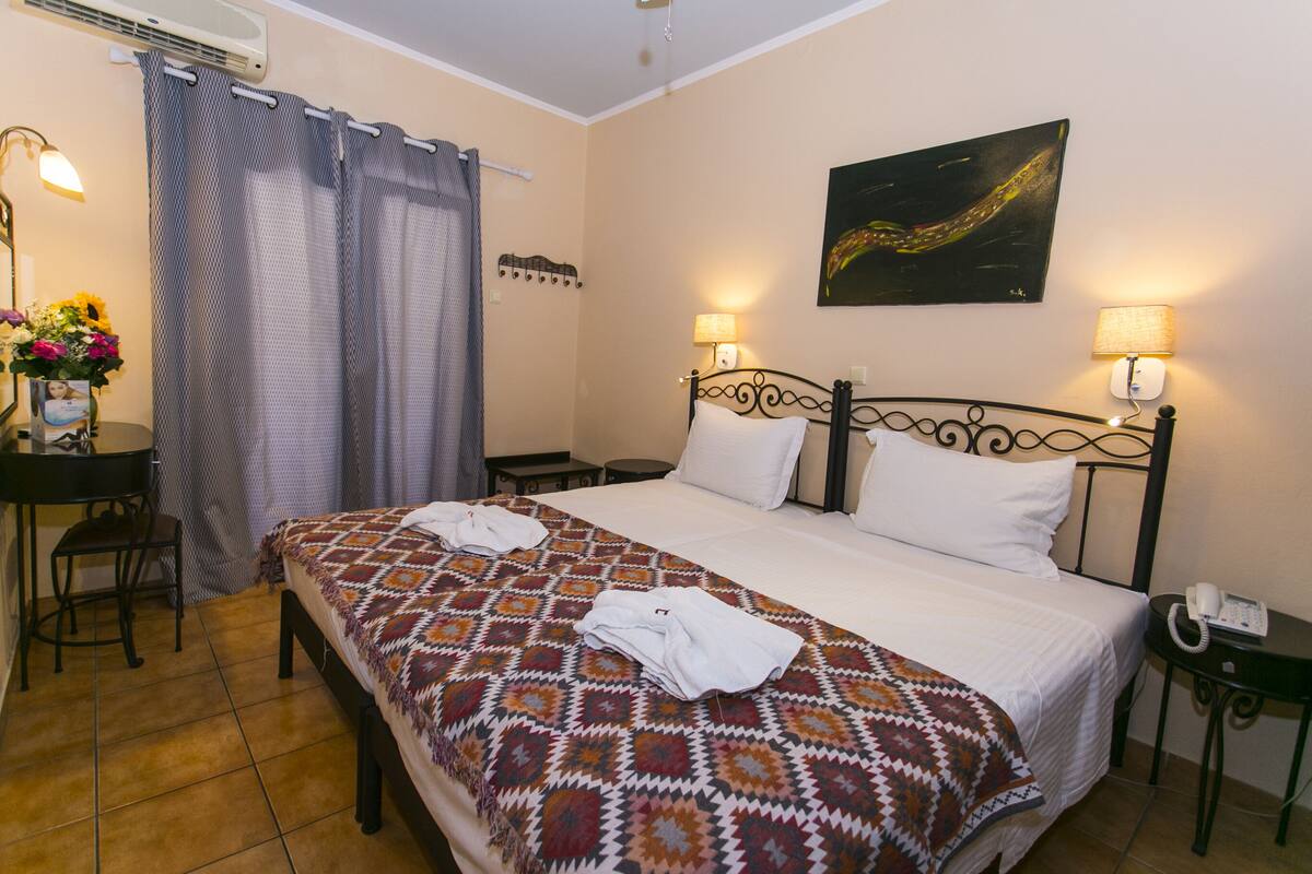 A hotel room with a bed, two bedside lamps, a desk with a chair, a small table, and a wall-mounted artwork.