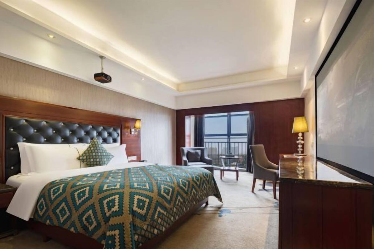 Habitación de hotel con una cama grande, cabecera de madera, balcón con una silla y escritorio con lámpara.