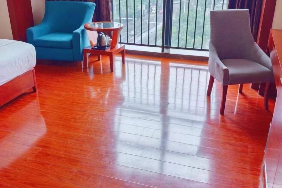 Habitación de hotel con suelo de madera, una cama, un sillón azul, un sillón gris y una mesita redonda con un hervidor.