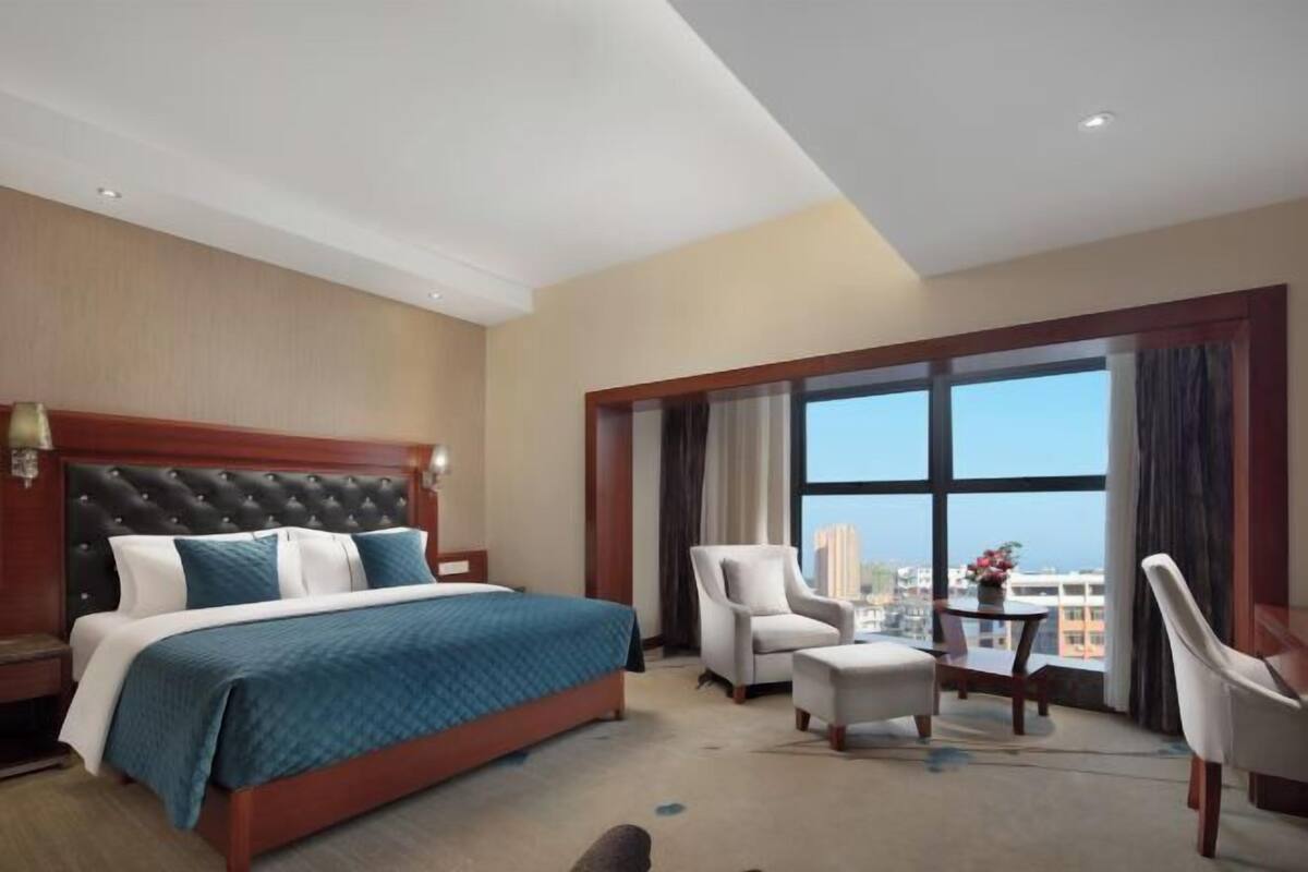 Habitación de hotel con una cama grande, zona de estar con una mesa y sillas, y un ventanal con vistas a la ciudad.