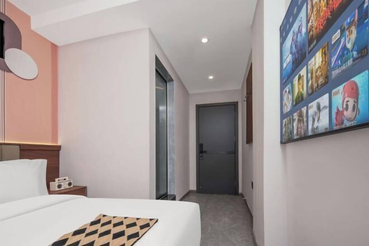 Una habitación de hotel con una cama, una mesita de noche, una pared con carteles y una puerta que da a otra habitación.