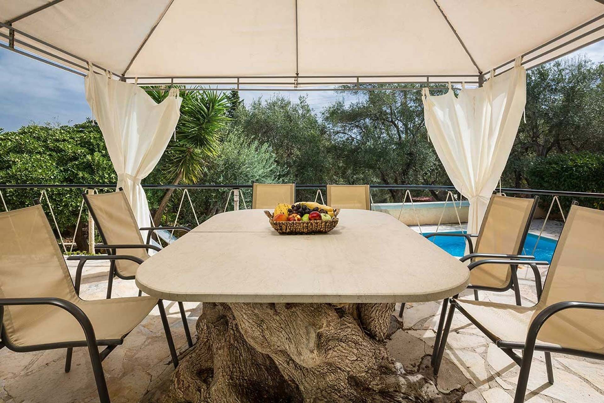 A covered outdoor dining area with a table and chairs, a fruit basket, and a pool in the background.