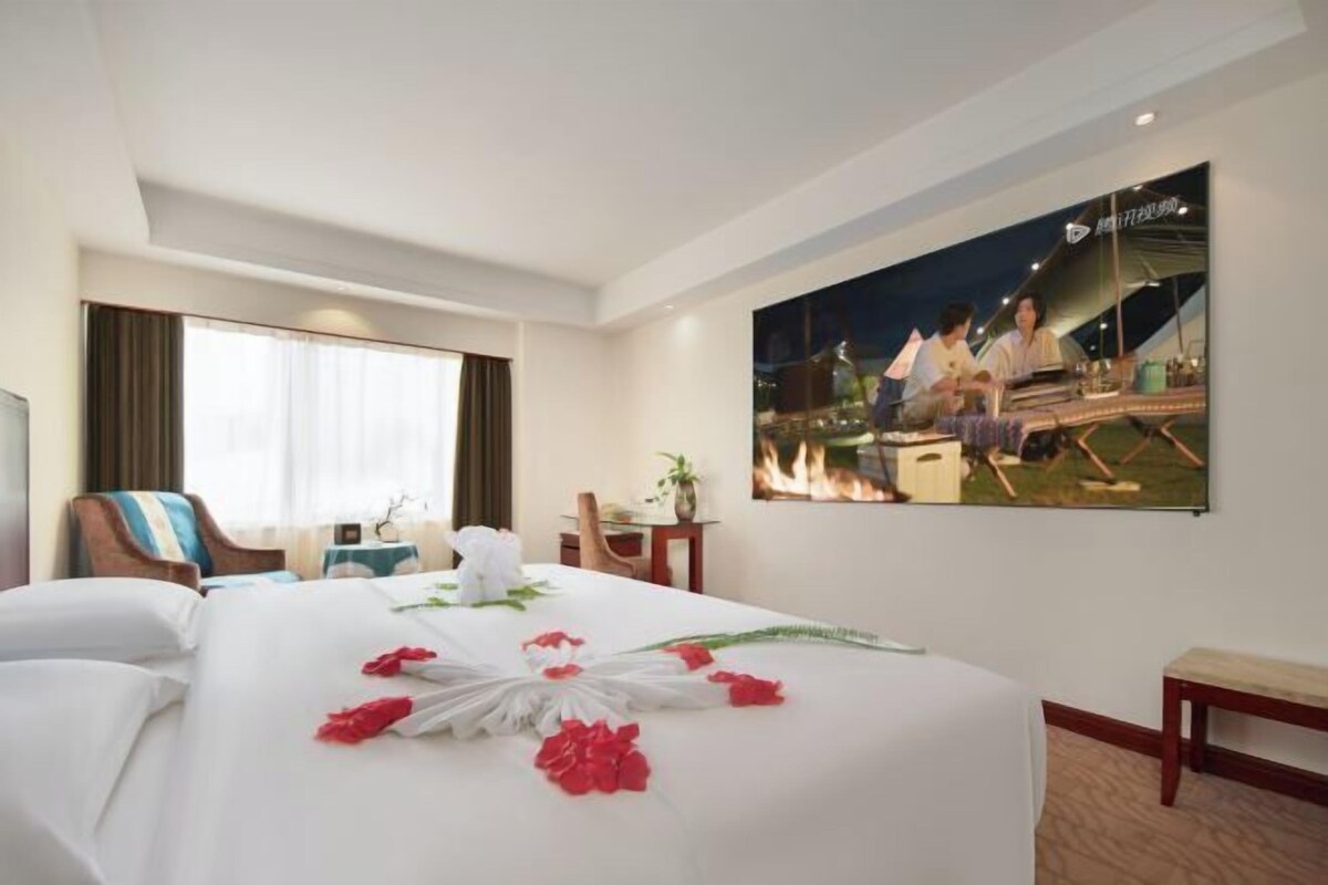 A hotel room with a large bed, a desk, a chair, a window with curtains, and a wall-mounted painting.
