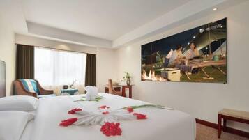 A hotel room with a large bed, a desk, a chair, a window with curtains, and a wall-mounted painting.