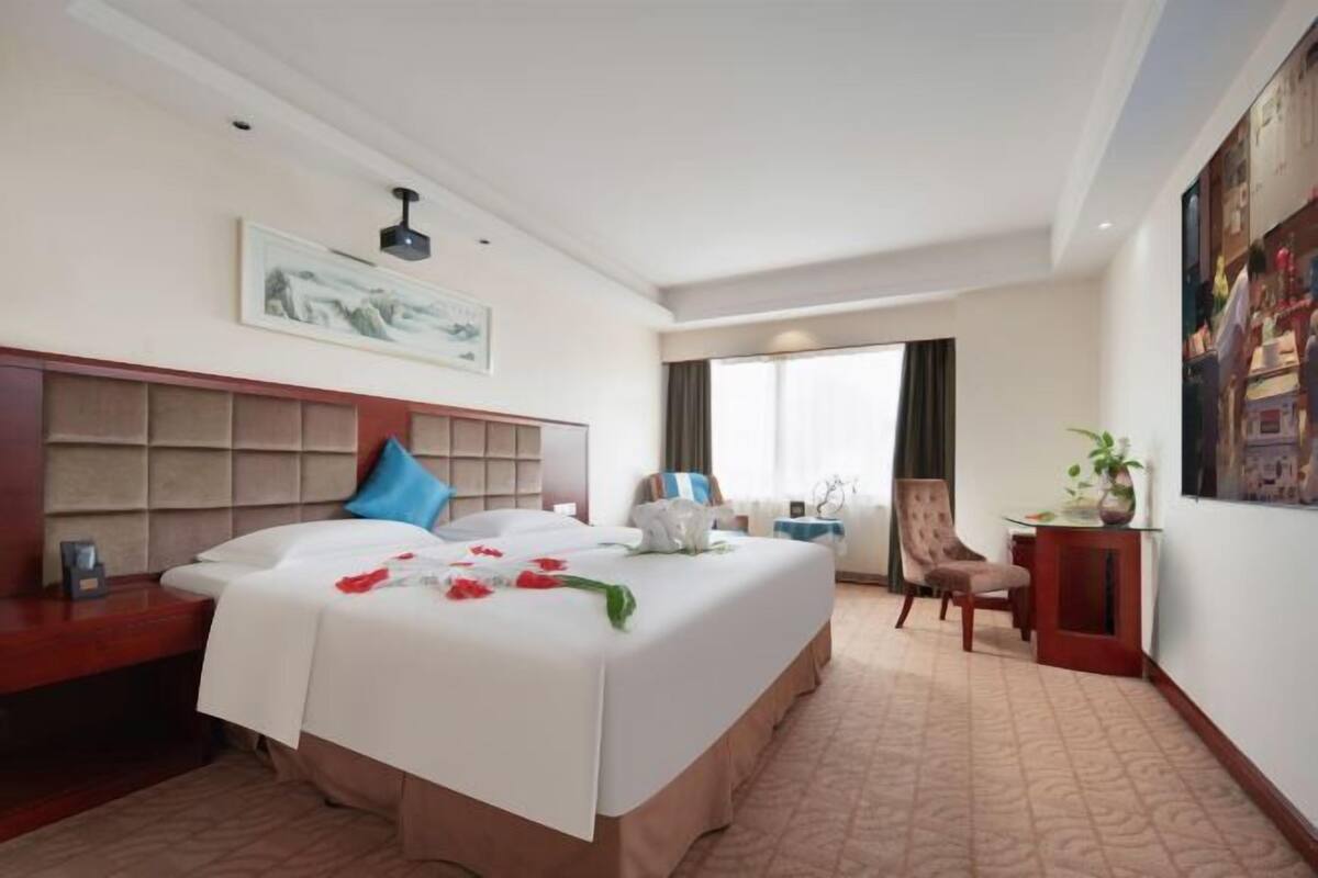 A hotel room with a large bed, a headboard, a bedside table, a chair, a desk, a window, and a painting on the wall.