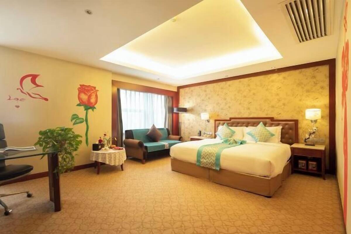 A hotel room with a bed, a desk, a chair, a small table with a plant, and a window with curtains.