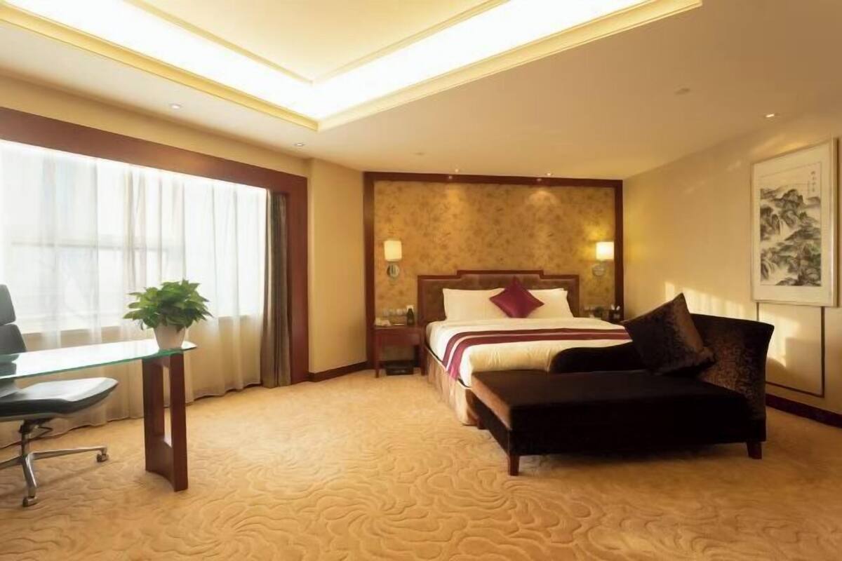 A hotel room with a bed, bedside table, lamp, chair, desk, and a potted plant.