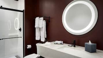Ein modernes Badezimmer mit Duschkabine aus Glas, einem runden Spiegel, einem weißen Waschbecken mit schwarzem Wasserhahn und einer Toilette.