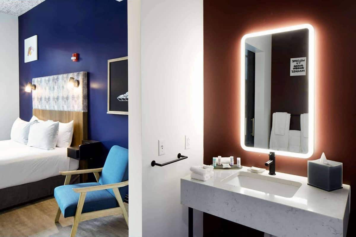 Ein Hotelzimmer mit Bett, blauem Sessel, Nachttisch, Wand-Fernseher und einem Badezimmer mit Waschbecken, Spiegel, Handtuchhalter und Toilettenartikeln.