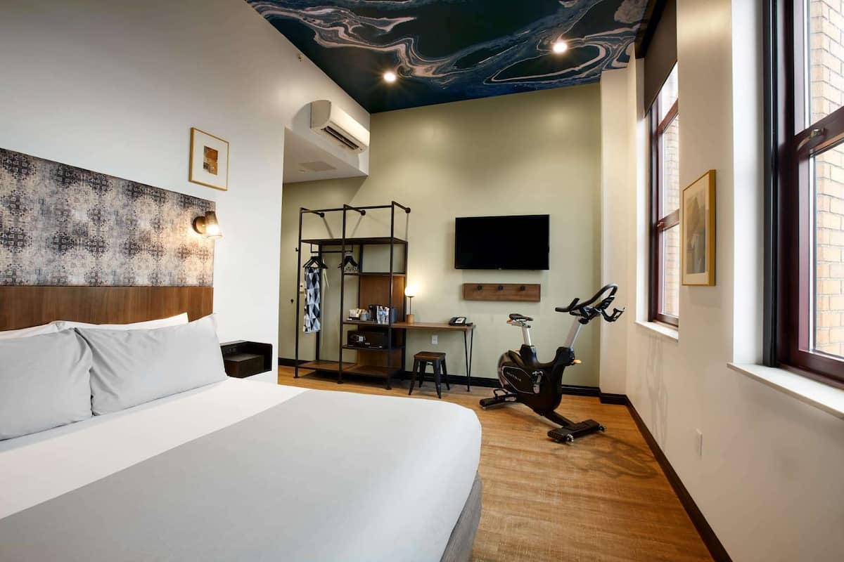 Ein modernes Hotelzimmer mit einem großen Bett, einem Trainingsfahrrad, einem Fernseher und einer Decke mit einem marmorähnlichen Design.