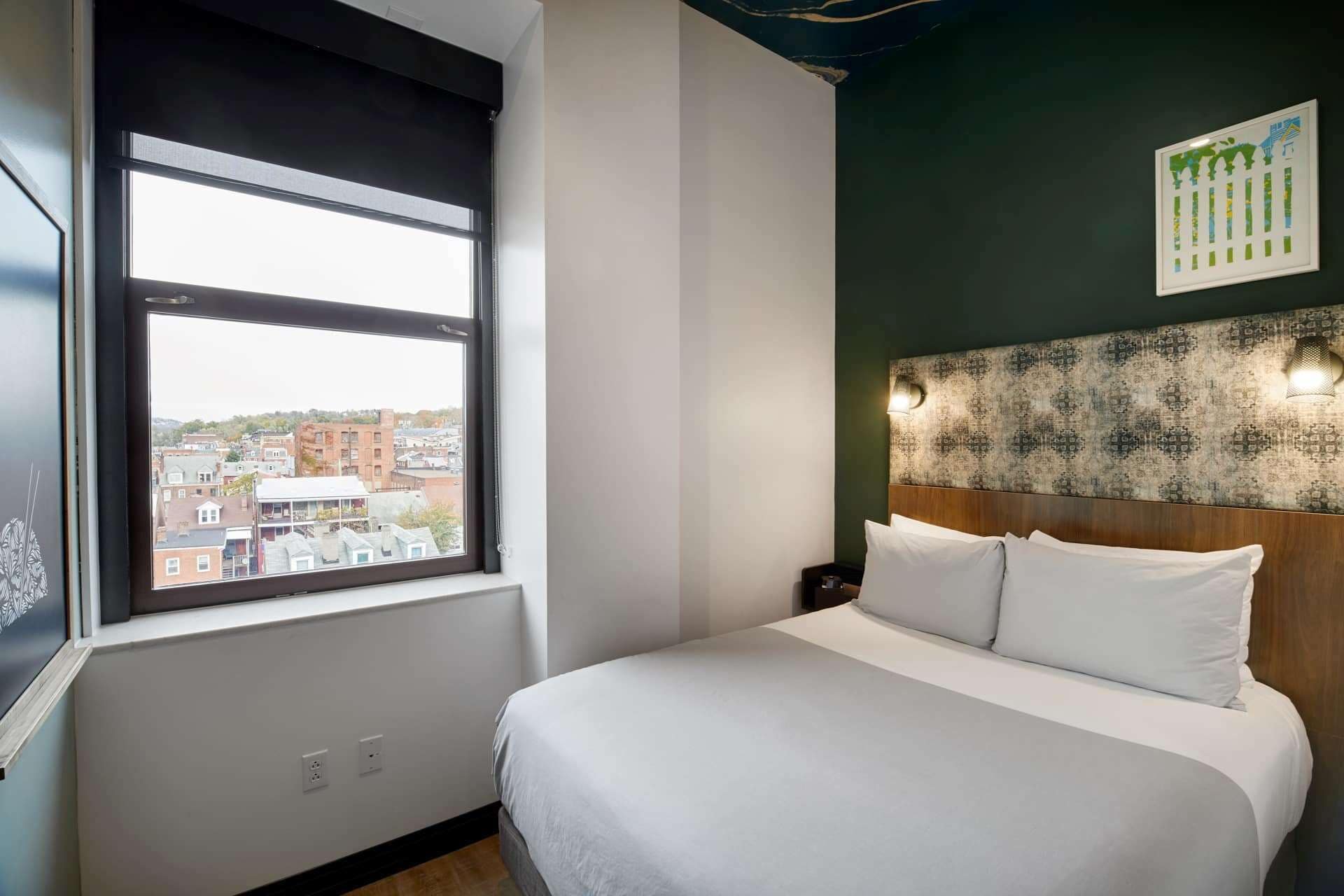 Une chambre d’hôtel dotée d’une grande fenêtre, d’un lit avec du linge de lit blanc, d’une tête de lit à motifs et d’un luminaire fixé au mur.