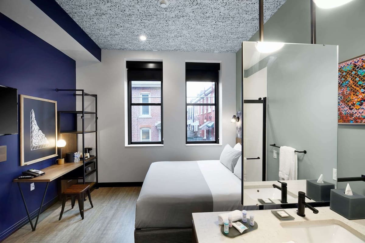 Ein modernes Hotelzimmer mit einem großen Bett, einem Schreibtisch mit Computer, einer Schreibtischlampe, zwei Fenstern mit Blick auf die Stadt, einem Badezimmer mit großem Spiegel und modernen Armaturen sowie einem farbenfrohen, abstrakten Gemälde an der Wand.
