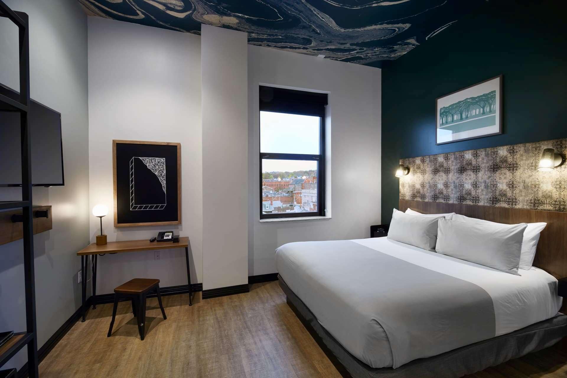 Habitación de hotel con una cama grande, un escritorio con una silla, un ventanal con vista a la ciudad y un techo con diseño que imita el mármol.