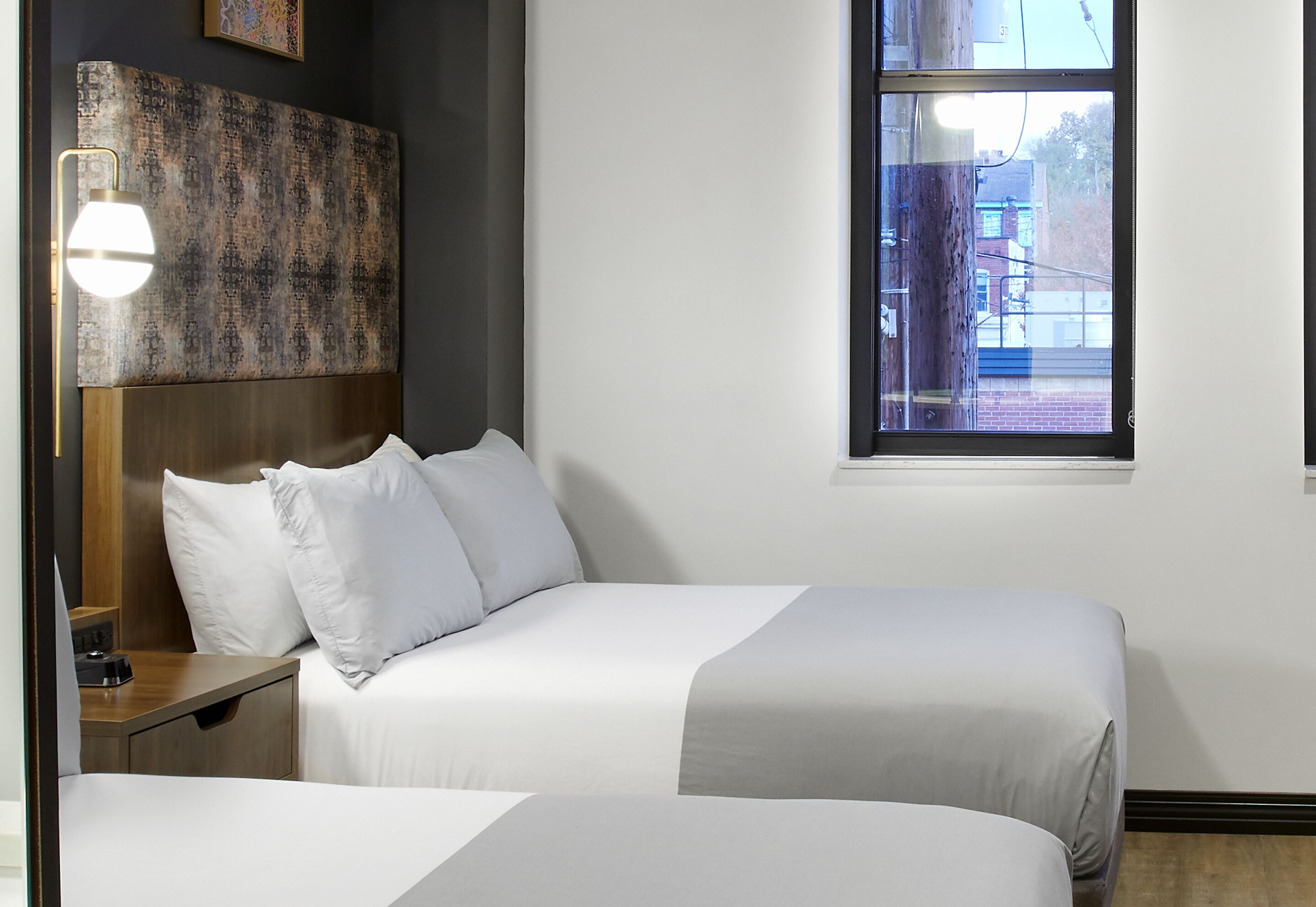 A hotel room with two beds, a headboard, a bedside table, a lamp, and a window with a view of buildings.