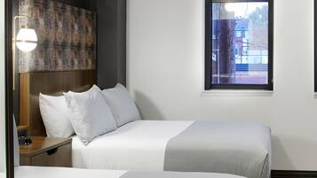 A hotel room with two beds, a headboard, a bedside table, a lamp, and a window with a view of buildings.