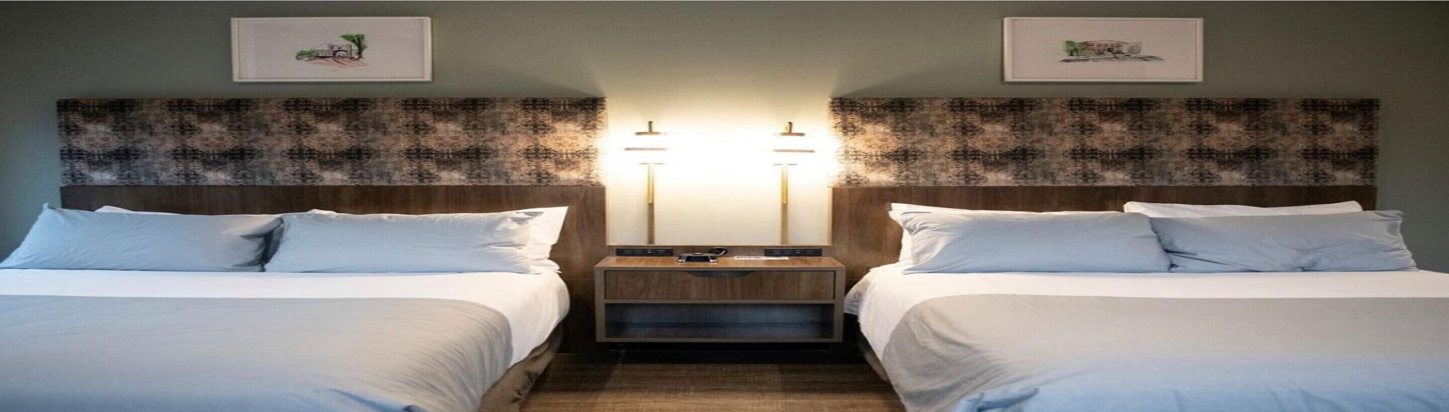 A hotel room with two beds, a nightstand with a lamp, and artwork on the walls.