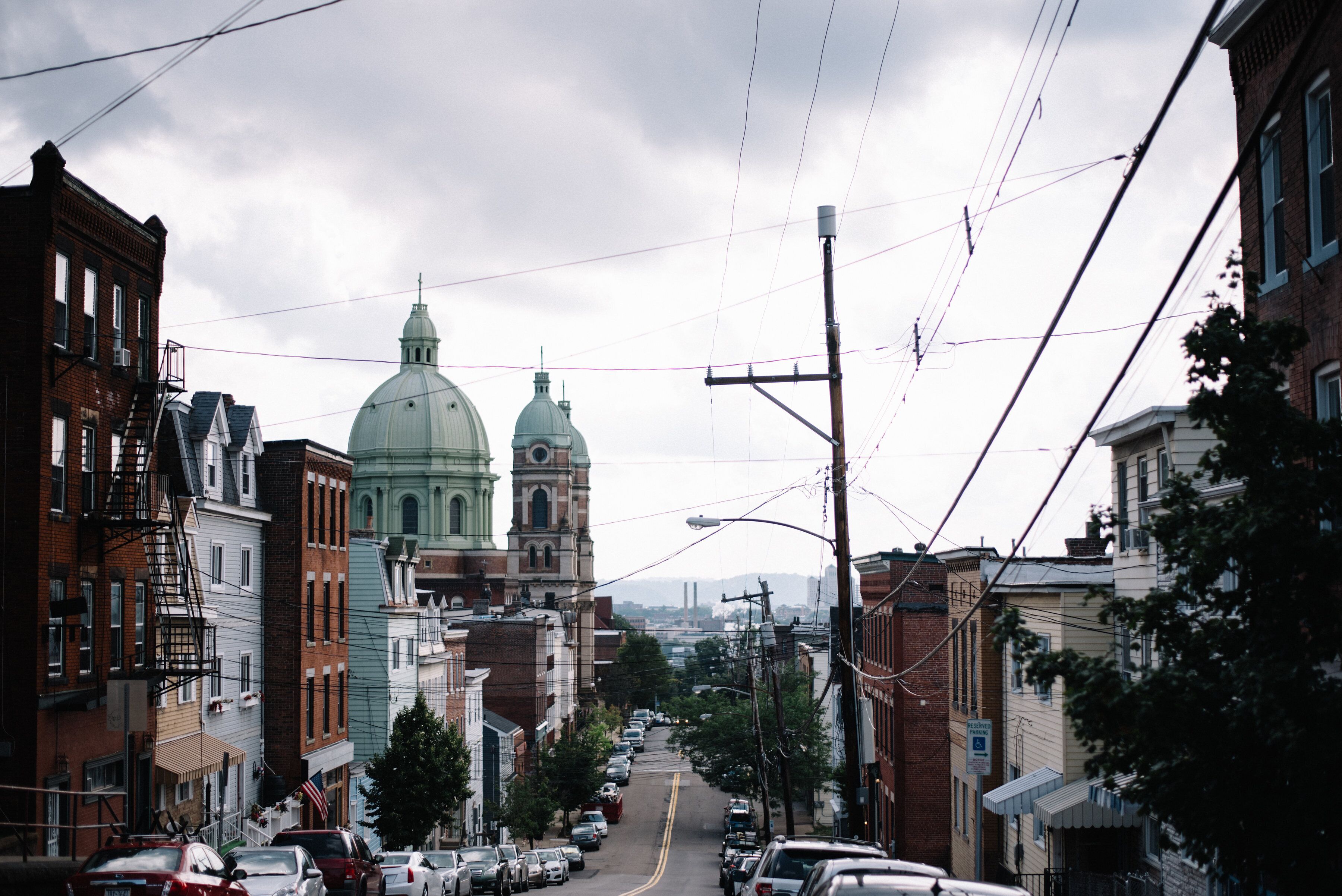 A street view with a large green dome building in the distance, surrounded by multi-story brick buildings with fire escapes.