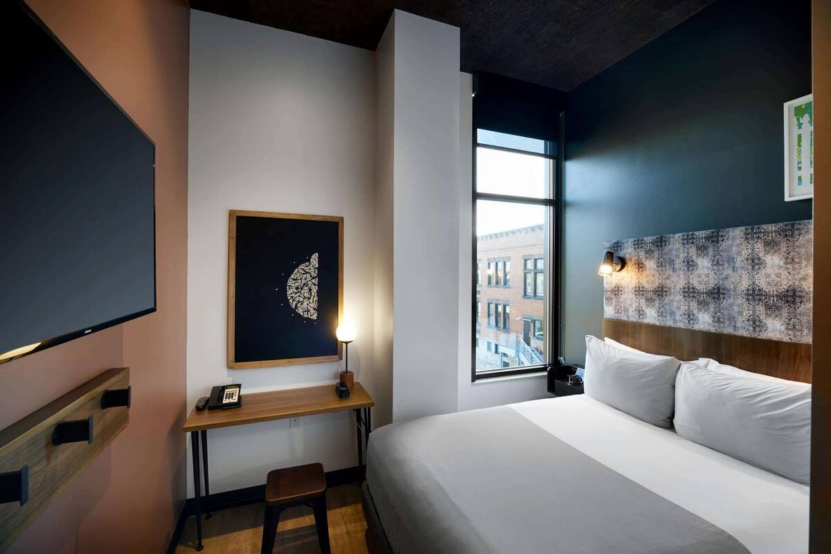 Ein modernes Hotelzimmer mit einem großen Bett, einem Schreibtisch mit Stuhl, einem Nachttisch, einem Wandbild und einem Fenster mit Blick auf Gebäude.