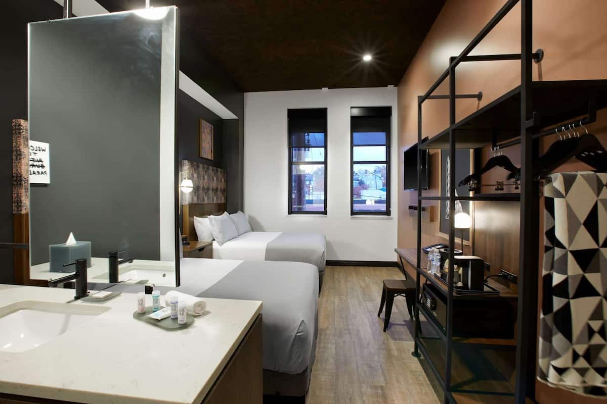 Ein Hotelzimmer mit einem großen Spiegel, einem Nachttisch mit Toilettenartikeln, zwei Betten, einem Kleiderschrank und Blick durch zwei Fenster nach draußen.