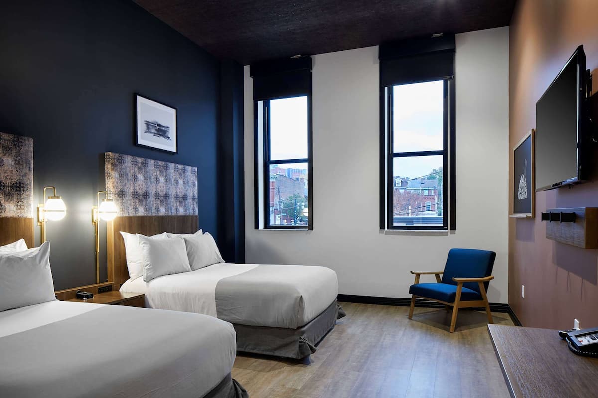 Ein Hotelzimmer mit zwei Betten, einem Sessel, einem Schreibtisch und großen Fenstern mit Blick auf die Stadt.