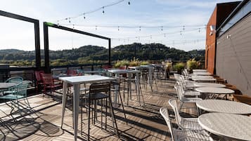 Een terras buiten met tafels en stoelen, lichtslingers en uitzicht op een beboste heuvel.