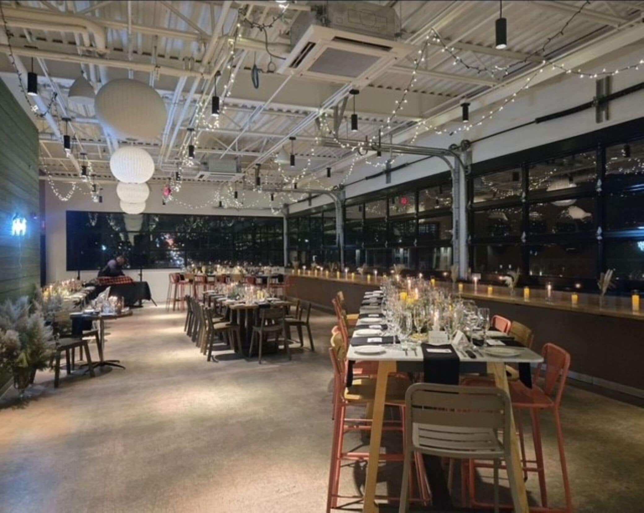 A restaurant with long tables set for dining, overhead lighting, and a view of the city through large windows.