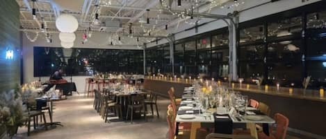 A restaurant with long tables set for dining, overhead lighting, and a view of the city through large windows.