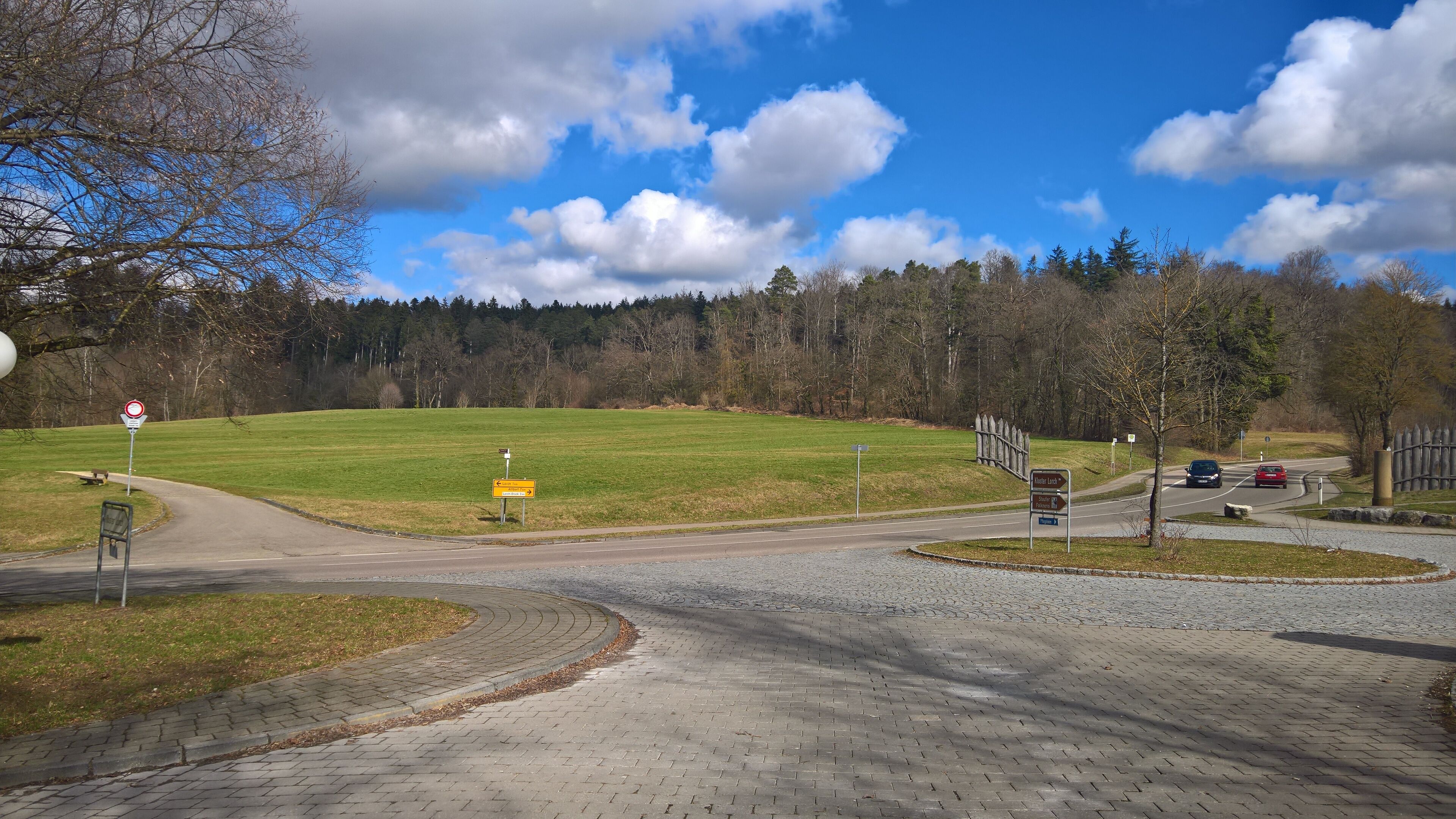 A paved intersection with directional signs, a grassy field, and a forest in the background.