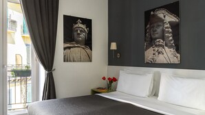 Habitación de hotel con cama, mesita de noche con un florero de flores rojas y dos cuadros de personajes históricos colgados en la pared.