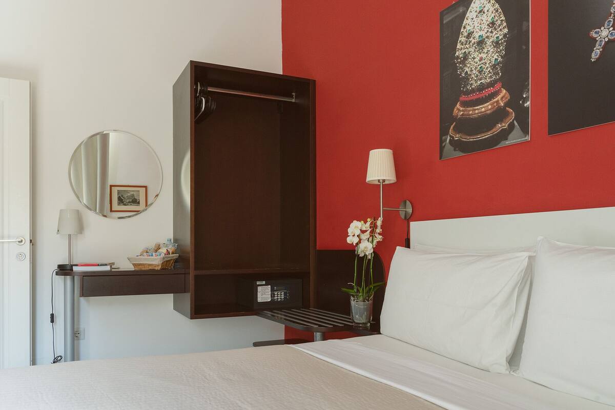 Habitación de hotel con cama, mesita de noche con lámpara, espejo y pared roja con obras de arte.