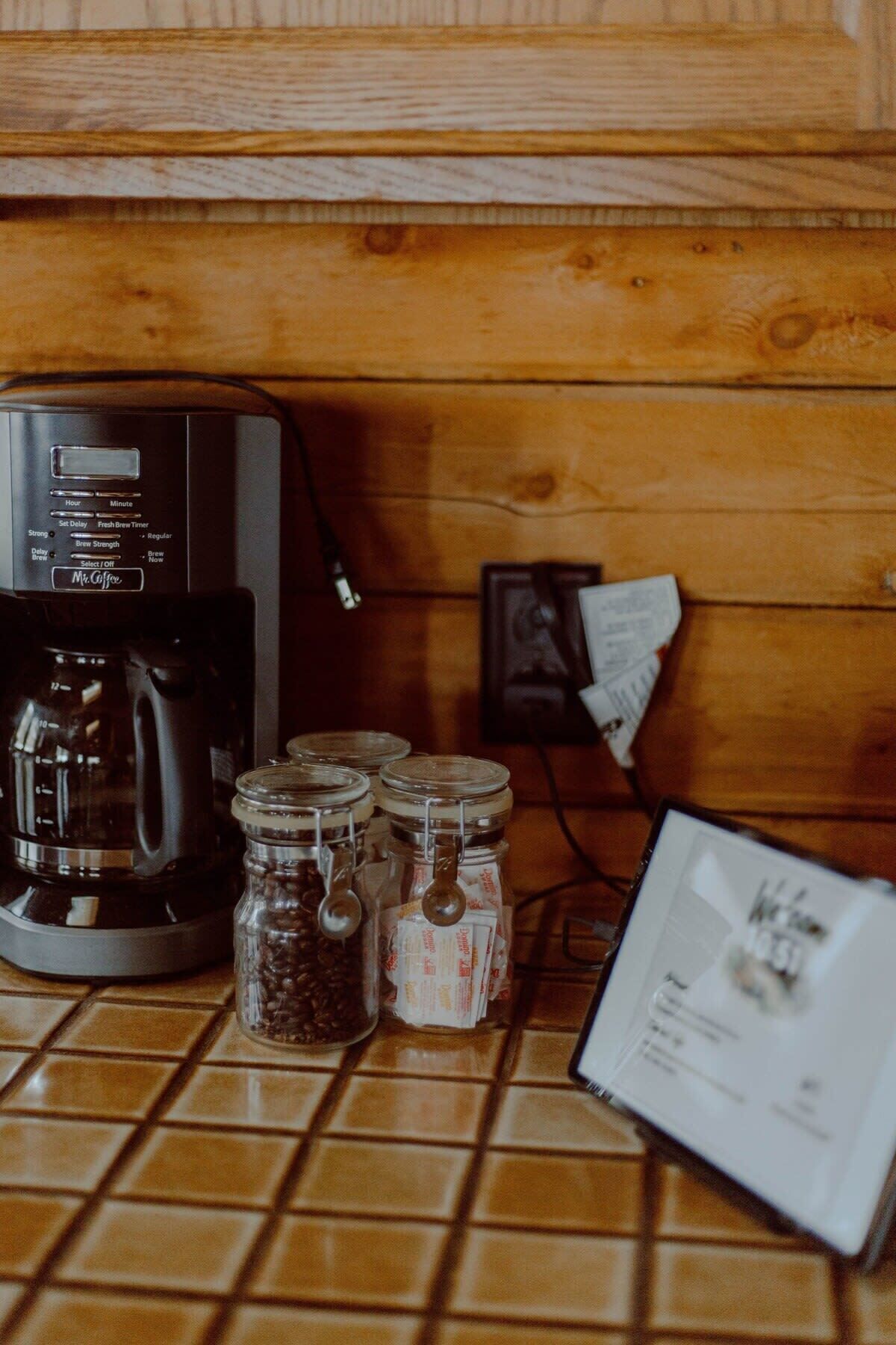 A coffee maker, jars of coffee beans, and a coffee bag on a tiled countertop.
